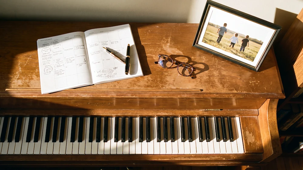 The Legacy Songbook open on a piano — planner pages, pen, reading glasses, and a family photo in warm afternoon light
